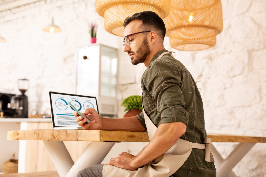 Bearded businessman reading message on phone with a laptop on his table showing his website's performance