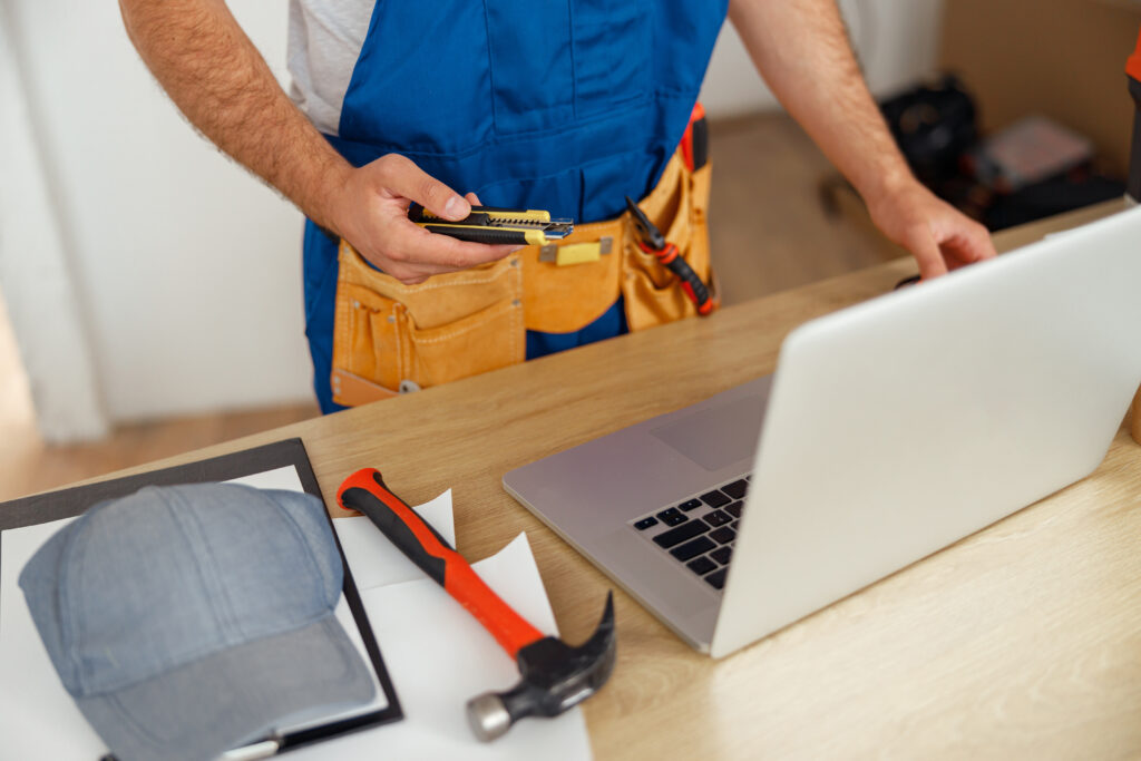 Cropped shot of repairman worker in uniform wearing tool belt, holding utitlity knife and using laptop while standing indoors. Occupation, home repair services concept